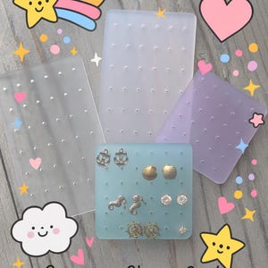 May include: A collection of earring display cards in clear, light blue, and lavender. The light blue card holds various stud earrings, including anchors, seashells, seahorses, and ship wheels. The background features decorative cartoon elements.