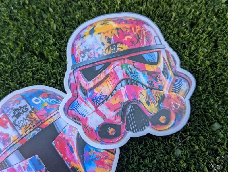 May include: A pair of colorful, graffiti-style Star Wars Stormtrooper helmet stickers. The helmets are painted in shades of pink, yellow, blue, and black.