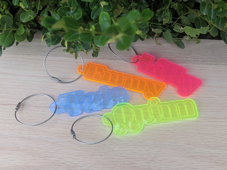 May include: Four neon-colored acrylic keychains with names "Lilly", "Emmy", "Quinn", and "Tyle" spelled out in a retro font. Each keychain is attached to a silver metal cable.