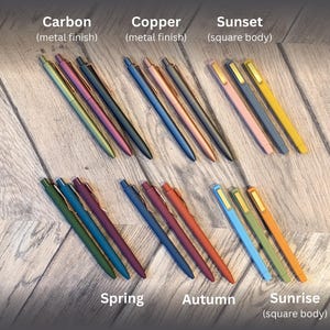 May include: A set of 3 packs of pens in various colors and finishes, including carbon, copper, sunset, spring, autumn, and sunrise. The pens are arranged in rows on a wooden surface.