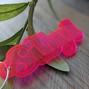 May include: A bright pink acrylic keychain with the word "silly" written in a playful, rounded font.