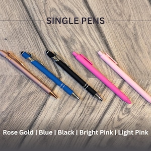 May include: Five pens on a wooden surface. The pens are rose gold, blue, black, bright pink, and light pink.  Single Pens.