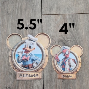 K&ouml;nnte beinhalten: Zwei runde Dekoschilder mit Micky-Maus-Ohren-Silhouette. Das gr&ouml;&szlig;ere 14 cm Schild zeigt Donald Duck und den Namen SHAUNA. Das kleinere 10 cm Schild zeigt Spider-Man und den Namen ISAIAH. Beide haben eine metallische Oberfl&auml;che.
