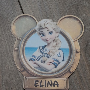 K&ouml;nnte beinhalten: Ein dekorativer Ausschnitt mit einem runden Bullaugenrahmen und Mickey-Mouse-Ohren. Im Bullauge befindet sich die Illustration einer blonden Figur in Matrosenkleidung vor einem Strandhintergrund. Ein rechteckiges Namensschild zeigt den Namen ELINA.