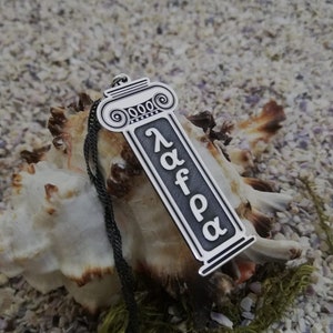 May include: Silver necklace with a pendant shaped like a column with the word "alpha" inscribed on it. The pendant is hanging from a black chain.