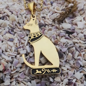 Pu&ograve; includere: Collana con ciondolo in oro con un designo di gatto egizio dettagliato con accenti neri e geroglifici. Il ciondolo &egrave; su una catena d'oro e riposa su un letto di conchiglie bianche.