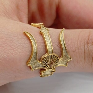 Pode incluir: Um anel dourado com um design de tridente. O tridente tem uma concha na base.