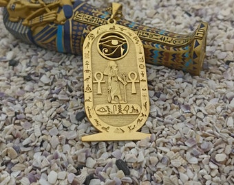 May include: Gold pendant necklace featuring an Egyptian Eye of Horus symbol, a standing figure of an Egyptian god, and hieroglyphics. The pendant is on a gold chain and is resting on a small, decorative, blue and gold sarcophagus.