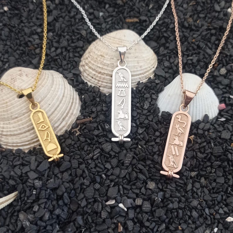 May include: Three gold, silver, and rose gold necklaces with Egyptian hieroglyph pendants. The pendants are rectangular and feature a design of a bird, a scarab beetle, and a sun disk.