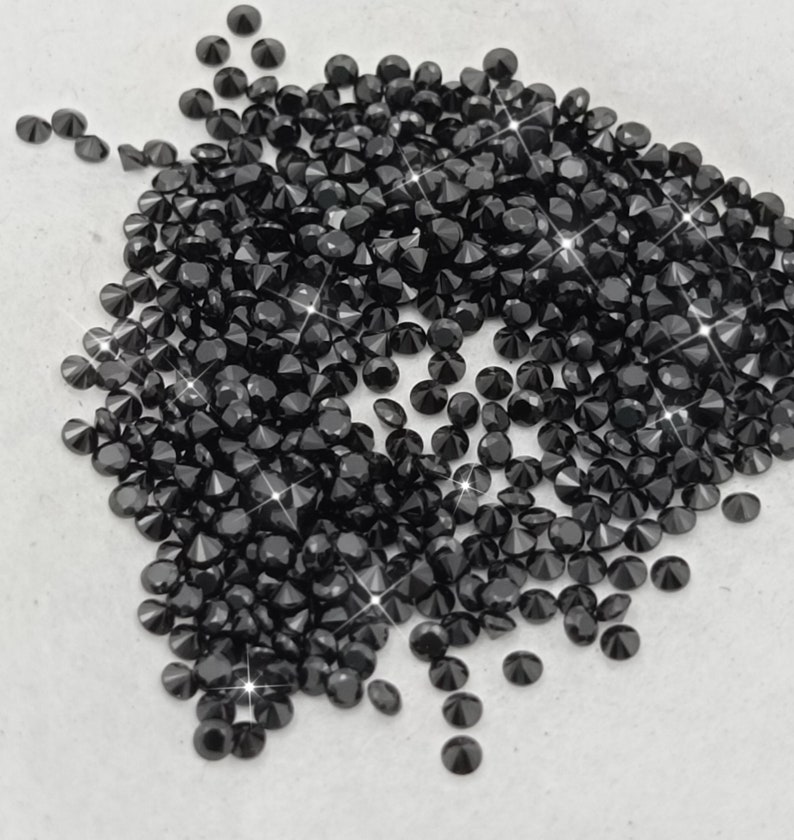 May include: A large pile of black diamond-shaped rhinestones. The rhinestones are scattered on a white surface.