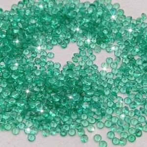 May include: A large pile of small, round, green gemstones. The gemstones are scattered on a white surface and have a shiny, reflective finish.