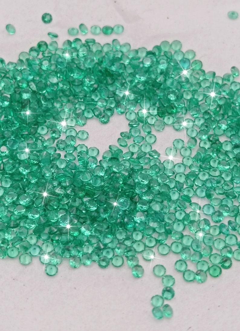 May include: A large pile of small, round, green gemstones. The gemstones are scattered on a white surface and have a shiny, reflective finish.