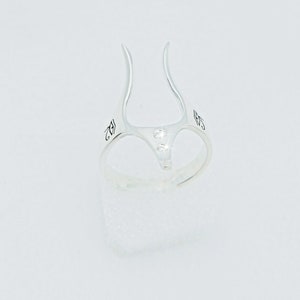 May include: Silver ring with two small clear stones set in the center. The ring has a unique design with two prongs that extend upwards.