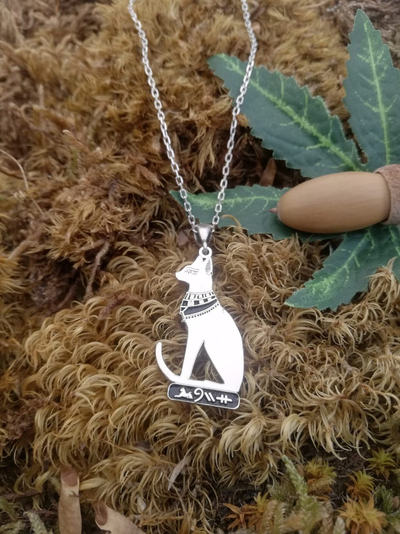 Pu&ograve; includere: Collana d'argento con un ciondolo a forma di gatto bianco con segni neri. Il gatto indossa un collare con un motivo bianco e nero. Il ciondolo ha anche geroglifici sul fondo.