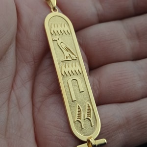 May include: Gold pendant necklace with an Egyptian hieroglyphic design featuring a bird, a symbol, and a pair of feathers.