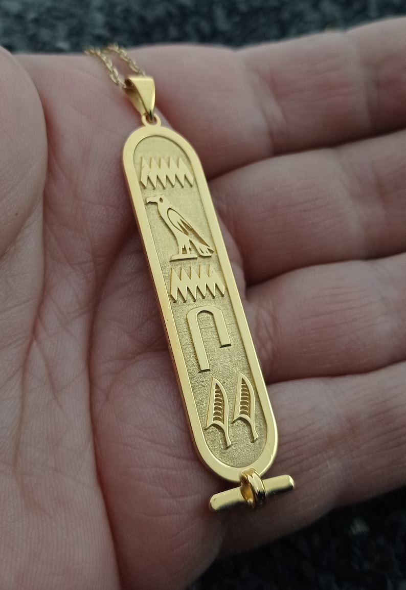 May include: Gold pendant necklace with an Egyptian hieroglyphic design featuring a bird, a symbol, and a pair of feathers.