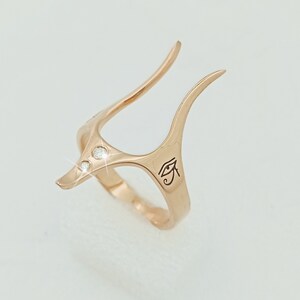 May include: A rose gold ring with a unique design featuring two prongs and a small eye of Horus symbol engraved on the band. The ring is adorned with three small clear gemstones.