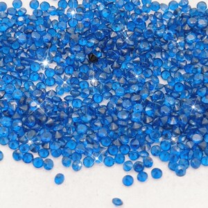 May include: A large pile of small, bright blue, faceted, round rhinestones. The rhinestones are scattered on a white surface.