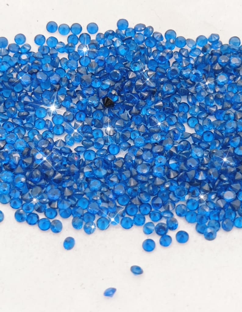 May include: A large pile of small, bright blue, faceted, round rhinestones. The rhinestones are scattered on a white surface.