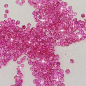May include: A large group of pink gemstones, likely rubies, are scattered on a white surface. The gemstones are round and have a faceted cut, reflecting light.