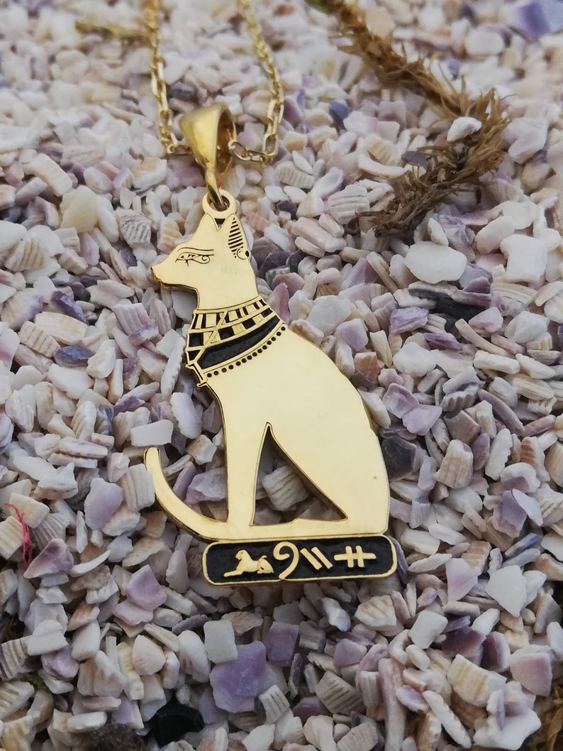 Pu&ograve; includere: Collana con ciondolo in oro con un designo di gatto egizio dettagliato con accenti neri e geroglifici. Il ciondolo &egrave; su una catena d'oro e riposa su un letto di conchiglie bianche.