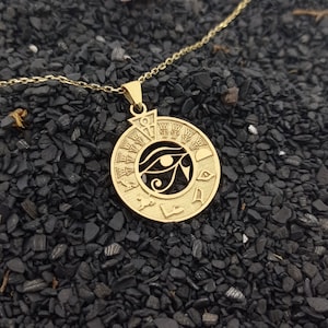 Könnte beinhalten: Goldkette mit einem runden Anhänger, der das Auge des Horus zeigt, ein altes ägyptisches Symbol für Schutz und Macht. Der Anhänger ist von Hieroglyphen umgeben.