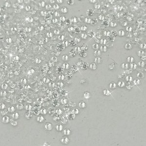 May include: A large quantity of clear, faceted rhinestones scattered on a white surface. The rhinestones are various sizes and shapes, creating a sparkling effect.