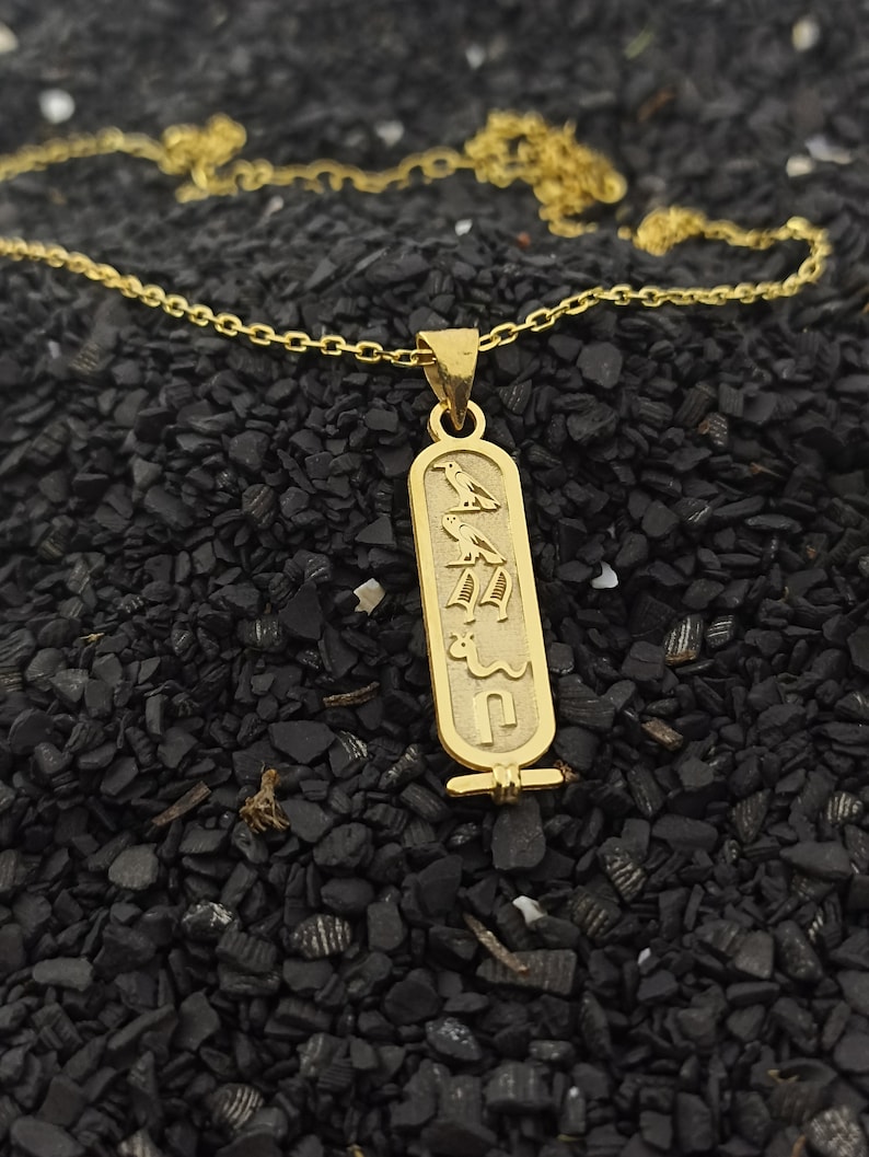 May include: A gold chain necklace with a pendant featuring an ancient Egyptian hieroglyphic design. The pendant is rectangular and has a detailed inscription.