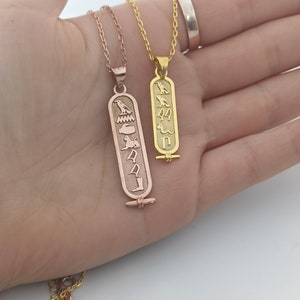 May include: Two gold necklaces with pendants featuring ancient Egyptian hieroglyphs. The pendant on the left is rose gold and the pendant on the right is yellow gold.