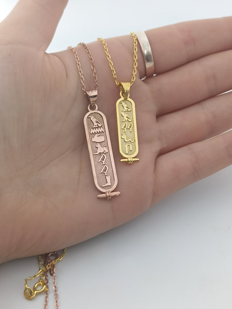 May include: Two gold necklaces with pendants featuring ancient Egyptian hieroglyphs. The pendant on the left is rose gold and the pendant on the right is yellow gold.