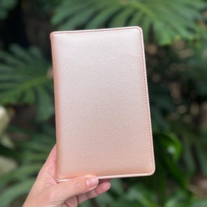 May include: A rose gold, rectangular journal or notebook with a textured surface. The cover is a solid color with a stitched border. The journal is held in a person's hand against a blurred green background.