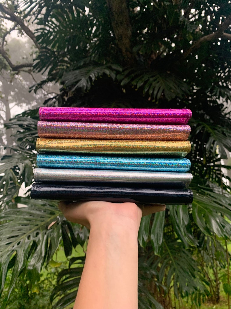 May include: A stack of six iridescent wallets in various colors, including pink, gold, blue, silver, and black. The wallets are made of a shiny, textured material.