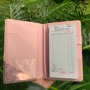 May include: A pink leather guest check folder with a clear plastic pocket on the front. The folder is open to reveal a white guest check with the text "Guest Check" and the number "8393-1".