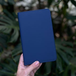 May include: A navy blue leather-textured passport holder. The cover is a solid, deep blue with a subtle grain. The edges are neatly stitched with a lighter blue thread. The holder is rectangular and held in a hand against a blurred green background.
