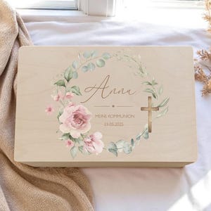 Puede incluir: Una caja de madera de color claro con un diseño de corona floral. La corona incluye rosas rosas, hojas verdes y una cruz dorada. El nombre "Anne" está escrito en escritura, junto con el texto "Meine Kommunion 23.05.2025".