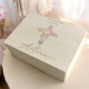 Puede incluir: Una caja de madera de color claro con un diseño de cruz floral. La caja tiene el nombre "Antonia" en escritura cursiva y el texto "ZUR KOMMUNION 24. Mai 2025". La caja es rectangular y parece ser de madera.
