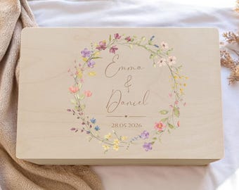 Op de afbeelding: Een lichtgekleurde houten kist met een bloemenkransontwerp. De krans heeft roze, paarse en gele bloemen. De namen "Emma & Daniel" en de datum "28.05.2026" staan in het midden geschreven.