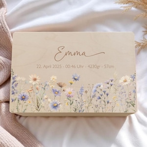 Può includere: Una scatola di legno color chiaro con un design floreale e il nome "Emma" in corsivo. La scatola riporta la data, l'ora, il peso (4230 grammi) e l'altezza (57 cm). Il design floreale presenta fiori blu, viola e bianchi.