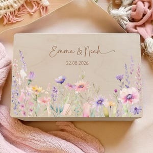 Op de afbeelding: Een houten kistje met een bloemenontwerp in roze, paarse en gele tinten. De kist is gepersonaliseerd met de namen "Emma & Noah" en de datum "22.08.2026" in sierlijke letters.