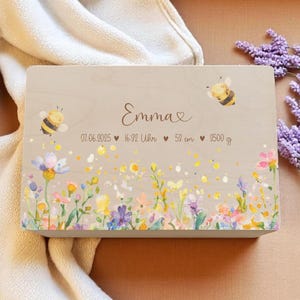 Può includere: Un cofanetto ricordo in legno chiaro con un design floreale e il nome "Emma". La scatola presenta illustrazioni di api e fiori. Il testo include la data 07.06.2025, l'ora, una lunghezza di 53 cm e un peso di 9500 g.