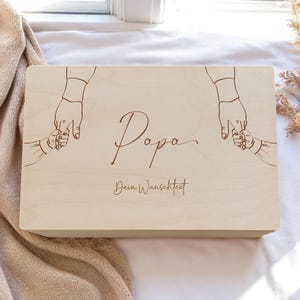Può includere: Una scatola di legno color chiaro con la parola "Papa" in corsivo. La scatola presenta un disegno inciso della mano di un genitore che tiene la mano di un bambino sulla parte superiore. Il testo "Dein Wunschtext" è anch'esso inciso sulla scatola.
