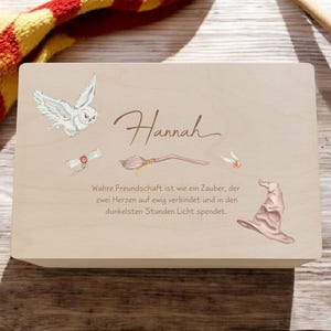 Peut inclure: Une boîte rectangulaire en bois clair portant le nom Hannah en écriture cursive, une chouette, un parchemin, un balai, un vif d'or et un chapeau de tri. Le texte indique : Wahre Freundschaft ist wie ein Zauber, der zwei Herzen auf ewig verbindet und in den dunkelsten Stunden Licht spendet.