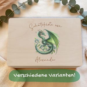 Op de afbeelding: Een lichtgekleurde houten kist met een groene drakenillustratie en de naam "Alexander". De kist heeft de tekst "Schatzkiste von" en "Verschiedene Varianten!". De kist staat op een beige ondergrond met decoratieve elementen.