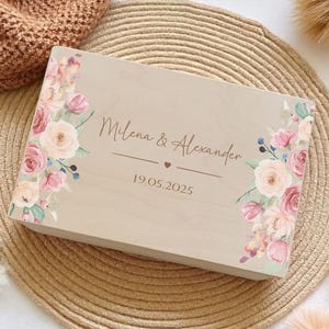 Puede incluir: Una caja rectangular de madera clara con un estampado floral de rosas rosas y crema en los lados izquierdo y derecho. El centro muestra el texto Milena & Alexander sobre un pequeño icono de corazón y la fecha 19.05.2025 en letra cursiva marrón.