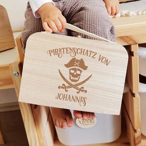 Könnte beinhalten: Eine hellfarbene Holzkiste mit Griff, mit einem Piratenmotiv. Das Design umfasst einen Totenkopf, gekreuzte Schwerter und den Text "Piratenschatz von Johannes". Die Kiste wird von einem Kind gehalten.