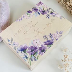 Könnte beinhalten: Eine rechteckige, helle Holzschatulle mit einem aufgedruckten Blumenmuster in Violett- und Blautönen. Auf dem Deckel befindet sich eine kursive Beschriftung: Die Liebe hört niemals auf, Rosemarie & Winfried, Simone, Josi, Anja, Jenna, Betsy, Nilla.
