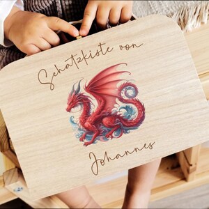 Puede incluir: Una caja de madera de color claro con una ilustración de un dragón rojo con detalles azules. El dragón tiene grandes alas y está rodeado de olas estilizadas. El texto "Schatzkiste von Johannes" está en cursiva.