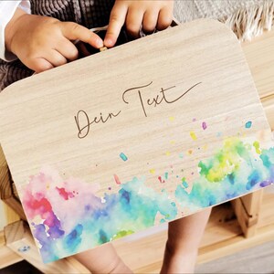 Könnte beinhalten: Eine Holzkiste mit hellem Holzmaserungsfinish. Die Kiste hat ein Aquarelldesign in Rosa-, Blau-, Grün- und Gelbtönen am unteren Rand. Der Text "Dein Text" ist in einer Schreibschrift auf der Oberseite der Kiste geschrieben.
