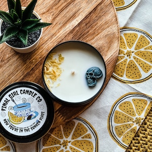 May include: A black candle with a white wax top and a blue skull on top. The candle is in a glass jar with a black lid. The candle is sitting on a wooden surface with a white and yellow patterned fabric underneath. The fabric has a lemon print. The candle jar has a label that says "Final Girl Candle Co. Now You're in the Sunken Place".