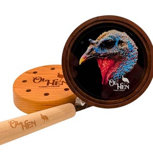 May include: A turkey call set featuring a round, wooden frame with a turkey head illustration. The set includes a wooden striker and a wooden sound board with the text "Ol' Hen Turkey Calls".